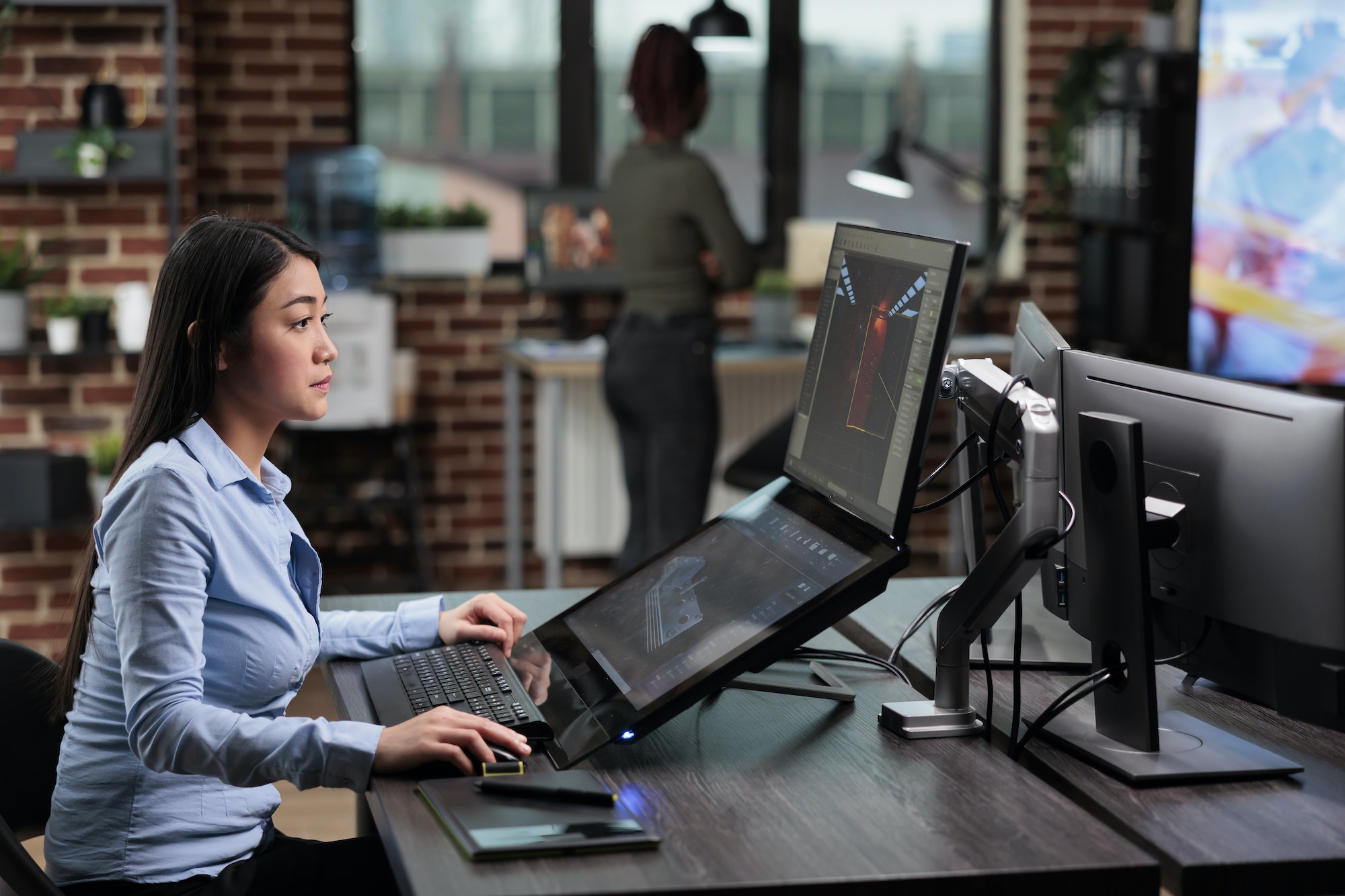 Confident asian 3D model creator sitting at desk with multiple monitors while working on CGI